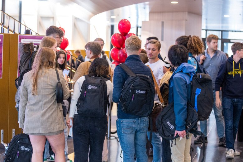 Students mingle in groups at the welcome fair with their KI backpacks. Balloons are visible in the background.