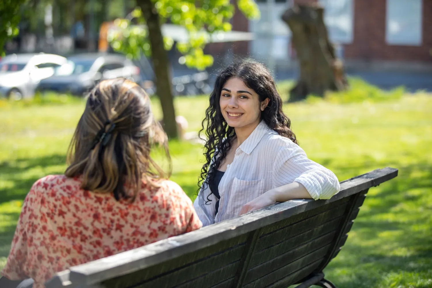 Students outside at campus Solna during a sunny spring day in 2022. || Two students sitting in the sun on a bench on campus. One looks into the camera and smiles.