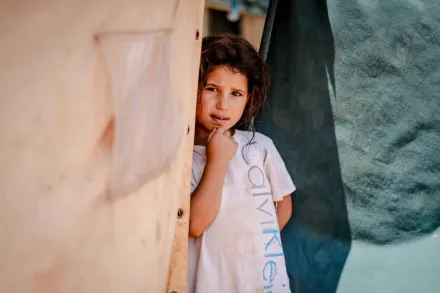 A girl in a refugee camp peers cautiously through the tent opening.